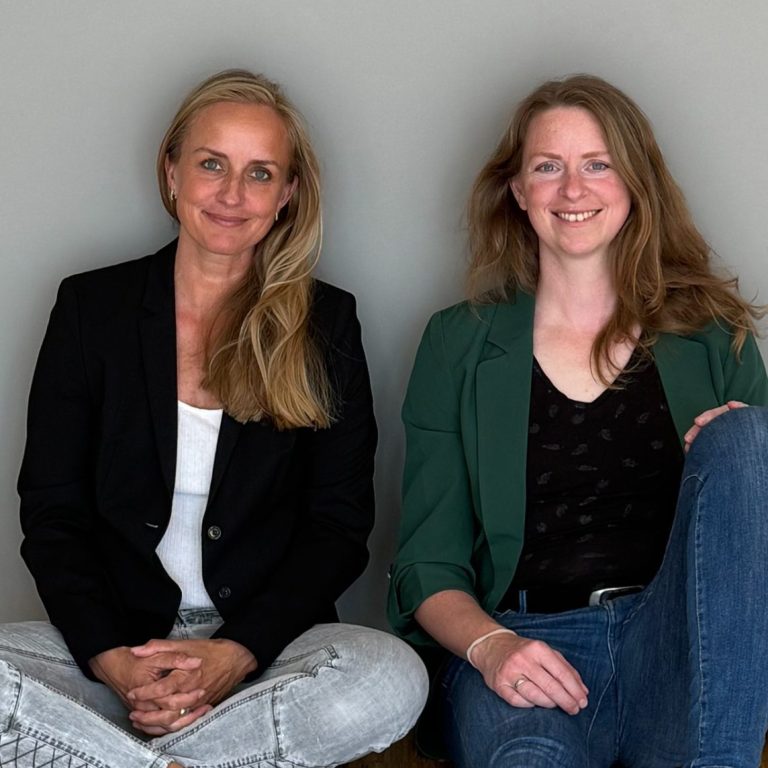 Elisa Wolff und Franziska Niedermeyer zwei Frauen stehen nebeneinander und lächeln einen an, links mit bunter Bluse, rechts wild print, freundlicher Blick, offene Haltung