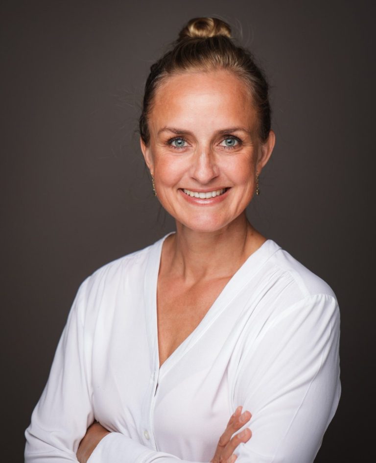 blonde Frau mit blauen Augen lächelt, Haare zusammengebunden, weißes Shirt, Arme locker verschränkt, blickt einen direkt an