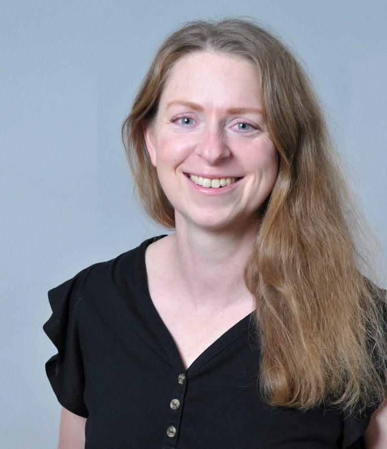 Blonde Frau, Haare schulterlang und offen, blaue Augen, lacht in die Kamera, schwarzes kurzärmeliges Shirt