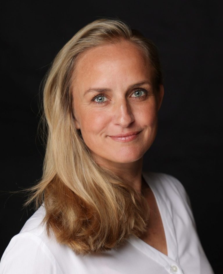 Foto von Elisa Wolff blonde Frau mit blauen Augen lächelt, Haare zusammengebunden, weißes Shirt, Arme locker verschränkt, blickt einen direkt an