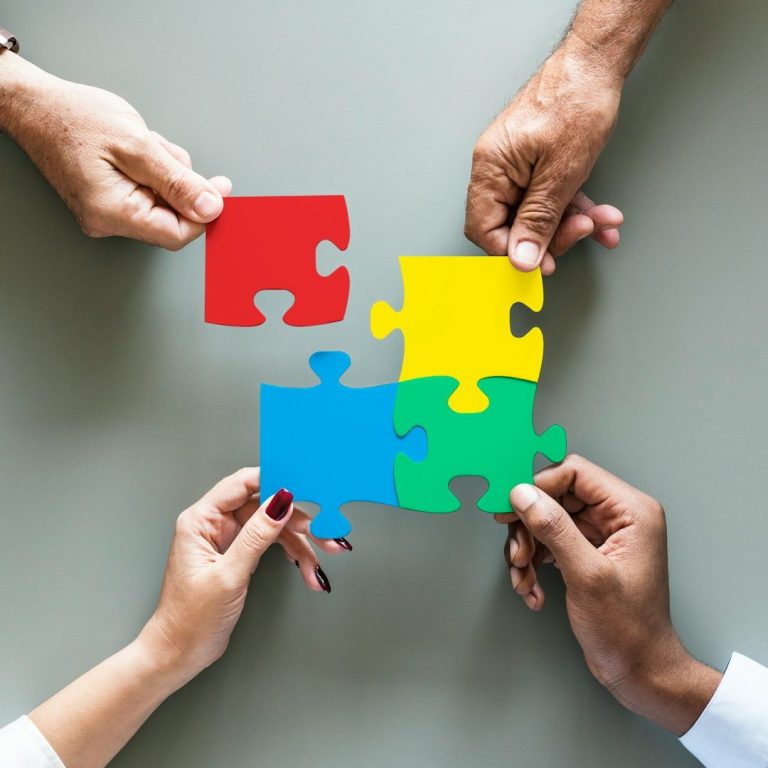 vier verschiedene Hände von Erwachsenen halten vier Puzzelteile in unterschiedlichen Farben (rot, gelb, blau und grün) zusammen.